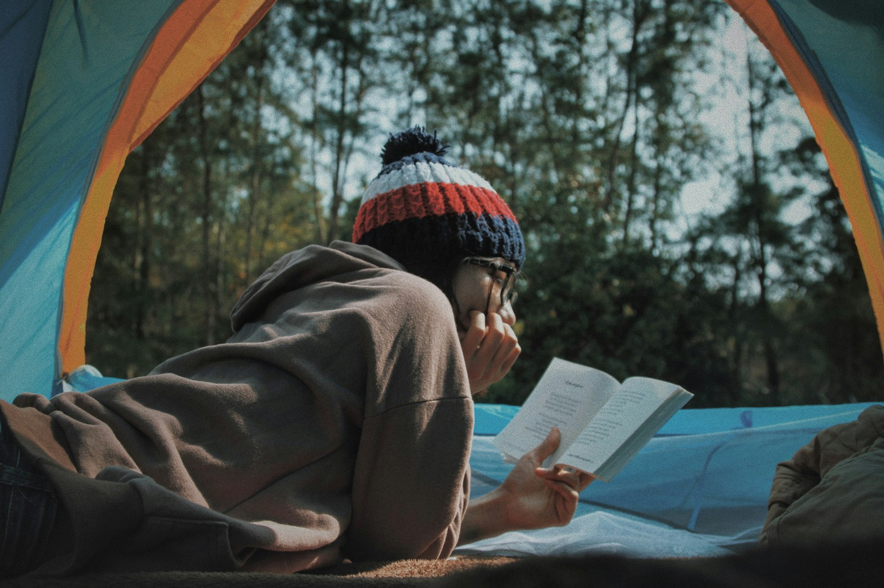 Pessoa deitada dentro de uma tenda aberta a ler um livro, com floresta ao fundo